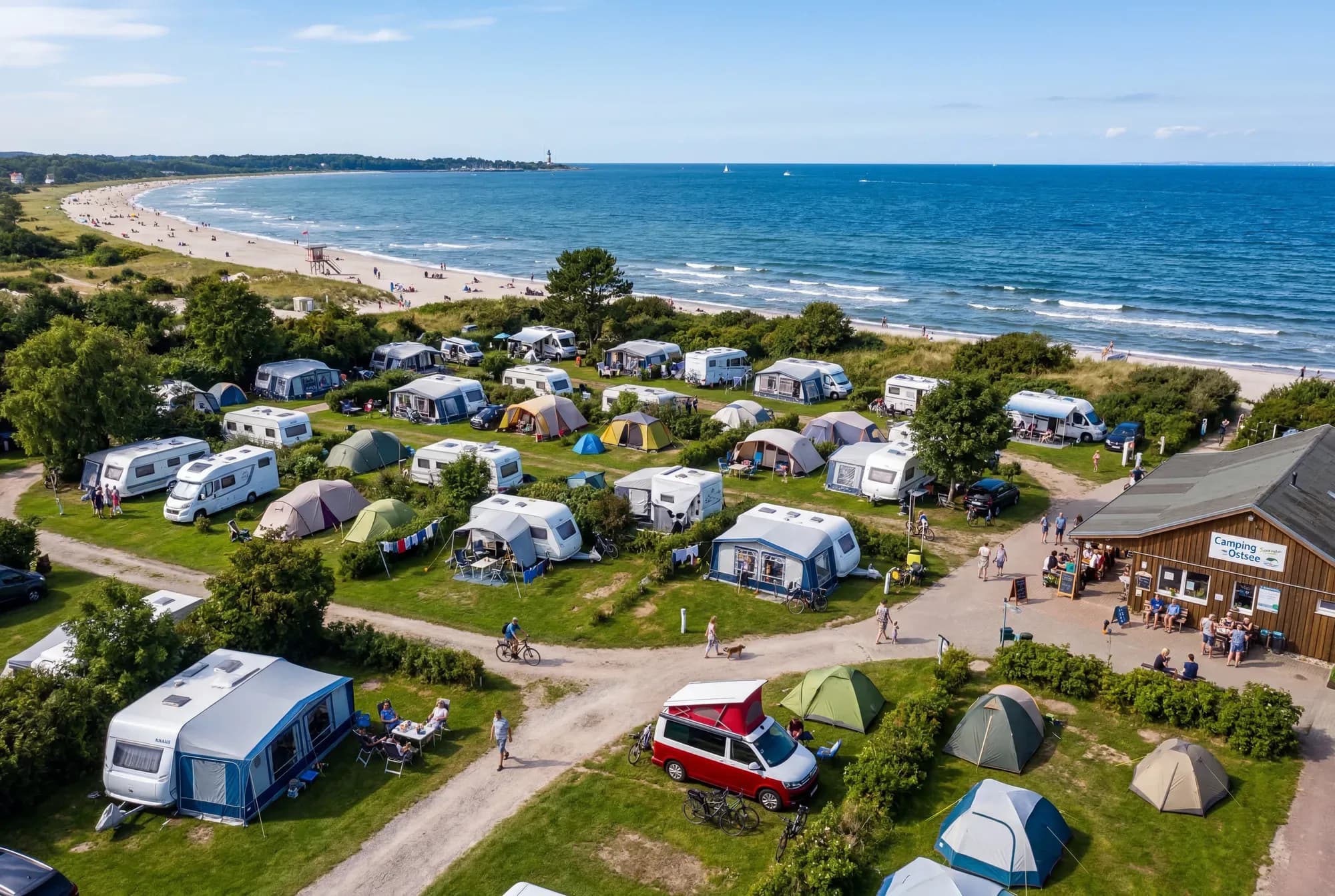 Luftaufnahme eines Campingplatzes an der Ostsee: Wohnwagen, Wohnmobile und Zelte auf Wiesen, dahinter Sandstrand und Meer