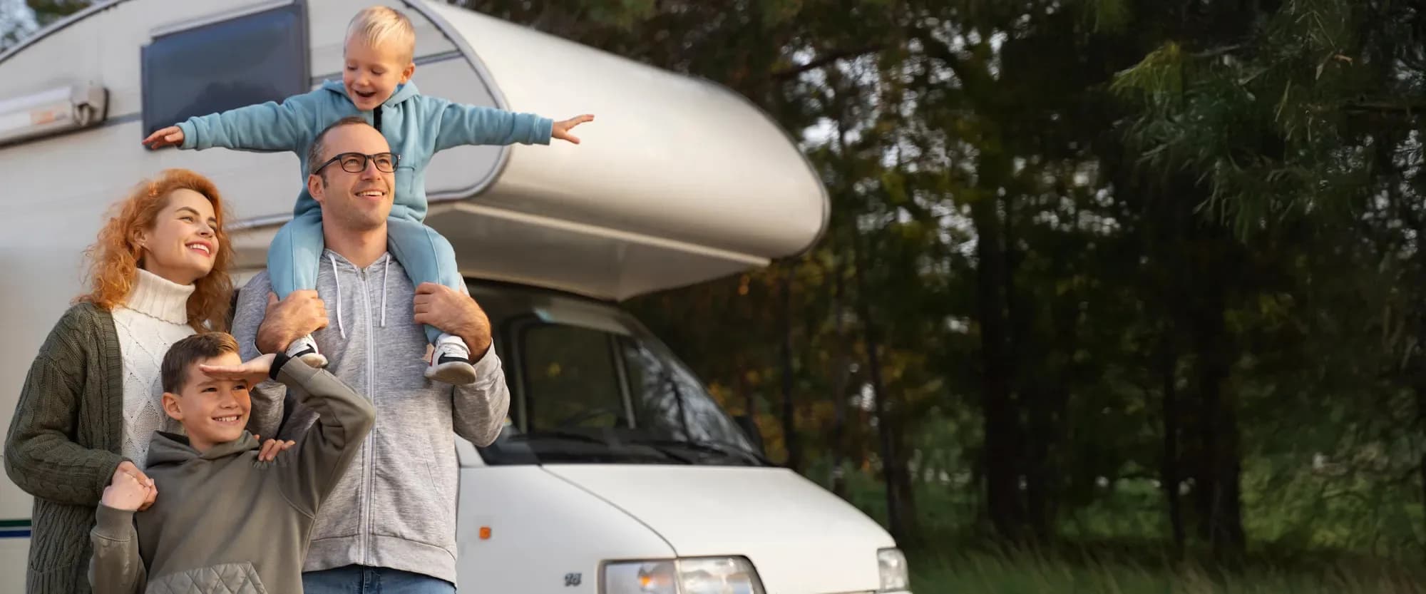 Familie mit zwei Kindern vor einem Wohnmobil am Waldrand, das jüngere Kind sitzt auf den Schultern des Vaters
